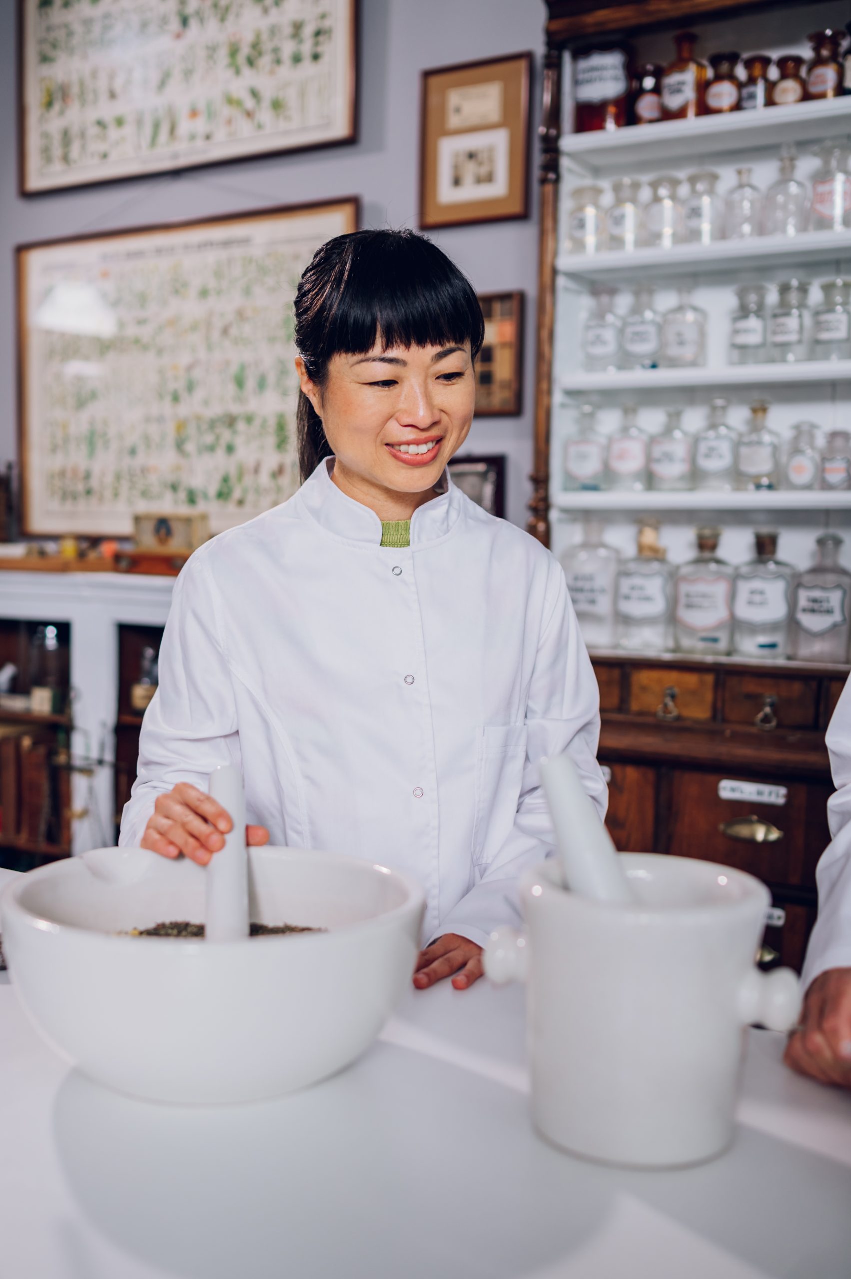 Une pharmacienne japonaise souriante en blouse de laboratoire se tient dans l'apothicaire avec son assistante et prépare des médicaments traditionnels dans un mortier. Médecine douce. Des collègues multiraciaux font un remède.<br />
